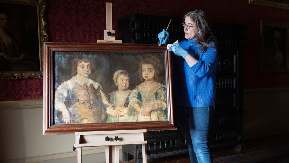 A curator brushing the frame of a print of the three eldest children of Charles I in a room with a red wall coverings