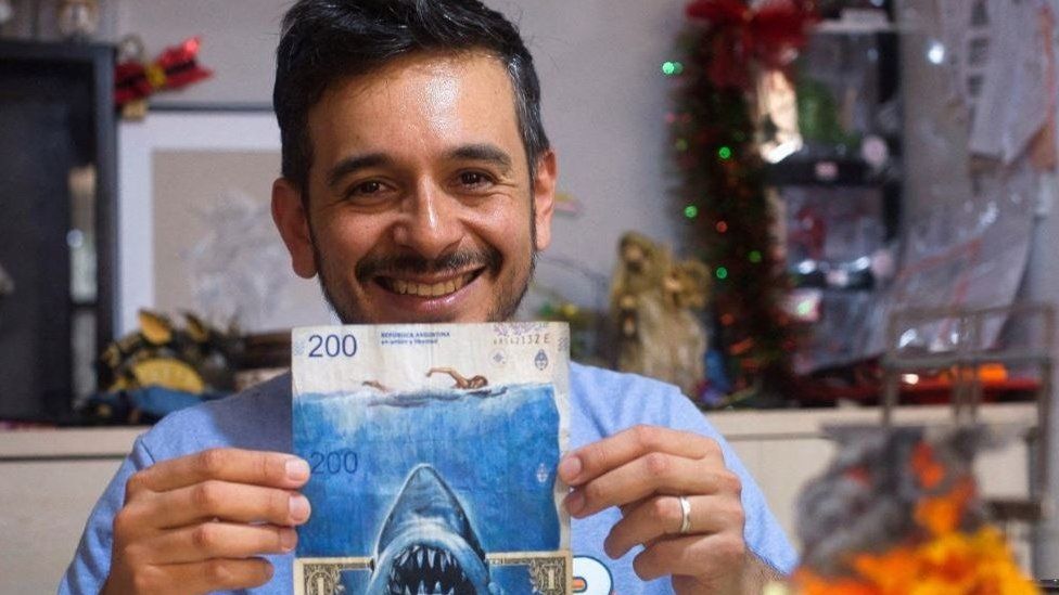 Artist Sergio Diaz holds intervened Argentine pesos bills and a US dollar depicting Steven Spielberg's movie "Jaws" as a parody of Argentina's ever-increasing inflation