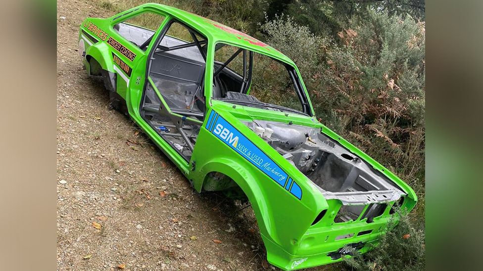 Green Ford rally car at the side of a gravel road stripped down the frame and body