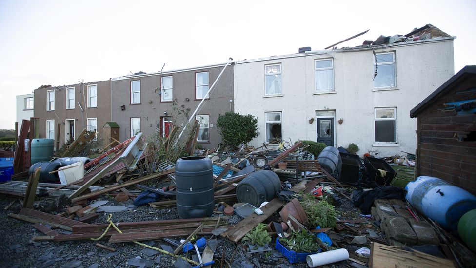 Photograph showing a row of houses with roofs ripped off and a lot of debris all over the front gardens.