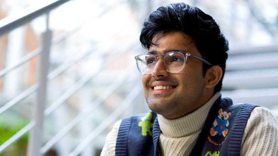 Sufiyaan Salam. Sufiyaan is a 26-year-old British Asian man. He has coiffed dark hair and wears clear-rimmed glasses. He's pictured sitting on stairs outside smiling to the right of the camera. He wears a navy knitted vest with koalas on it over a cream cable-knit high necked jumper.