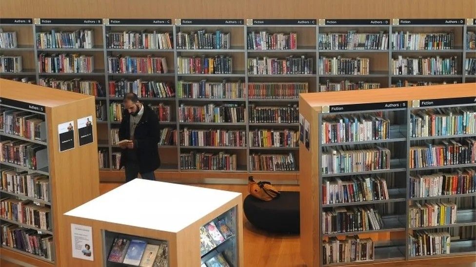 A man is standing reading a book in a library. He is surrounded by brown shelves filled with hundreds of colourful books 