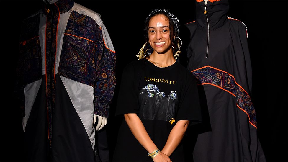 Kazna Asker during the Kazna Asker Presentation during London Fashion Week. Kazna is smiling wearing a dark head covering and a black top with the words "community" printed in gold and a design below it. Her hands are together. The background on either side has two of Kazna's designs, one is printed dark with white outline. The other on the right is dark with a printed pattern running across the middle.