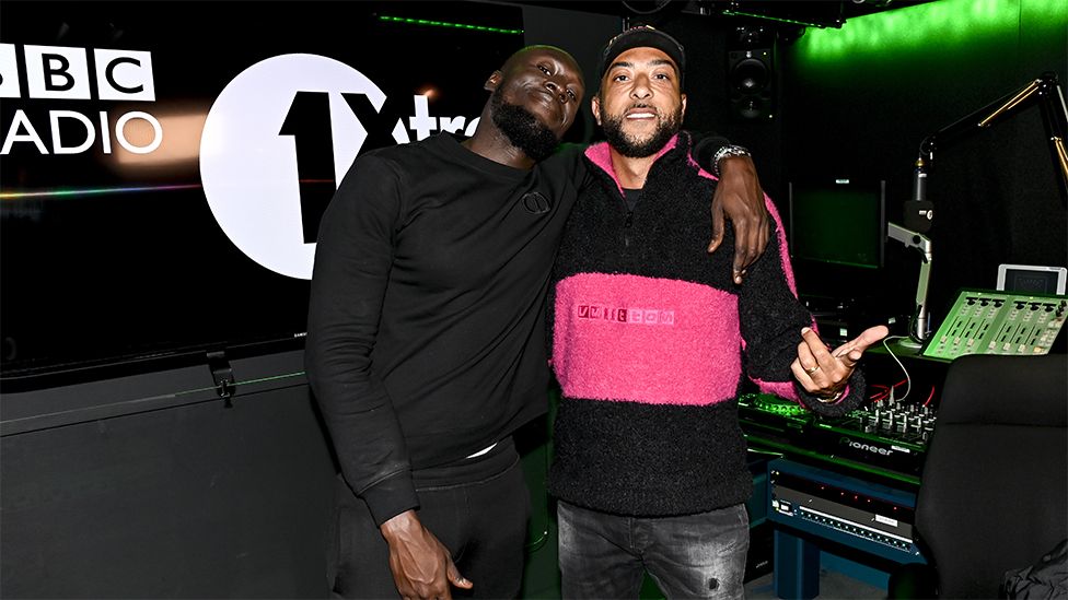 Stormzy and DJ Target in BBC 1Xtra studio. Stormzy is on the left wearing a black long sleeved top with his arm around Target, who is wearing a black and pink zipped jumper. Target's left hand is pointing towards the camera. In the background there is branding on a black background of "BBC Radio 1Xtra", green neon lights and a radio fader board with lots of buttons.