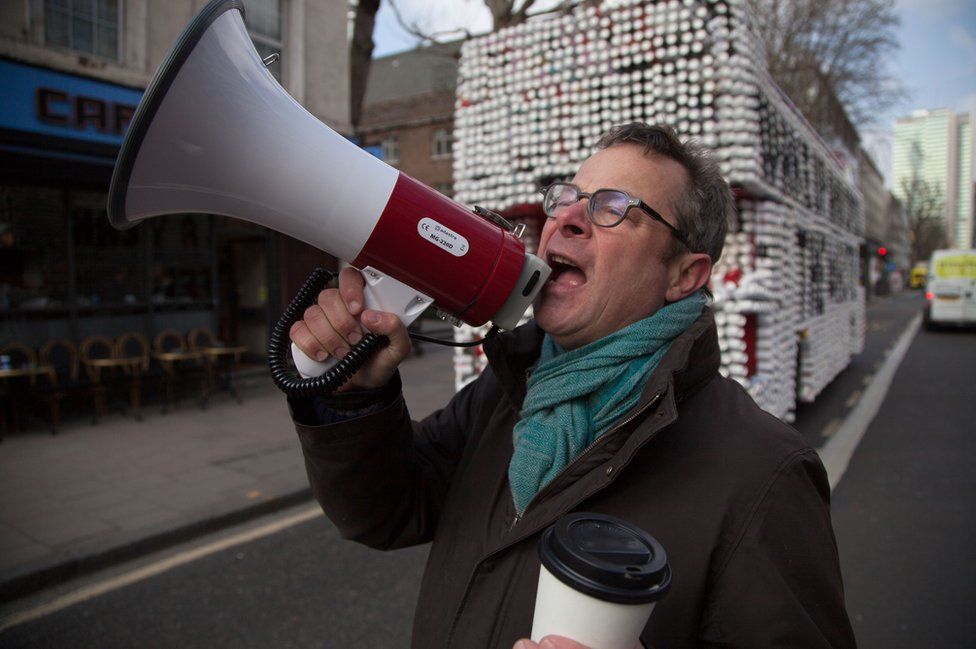 Hugh Fearnley-Whittingstall standing with a megaphone in front of a bus covered cups