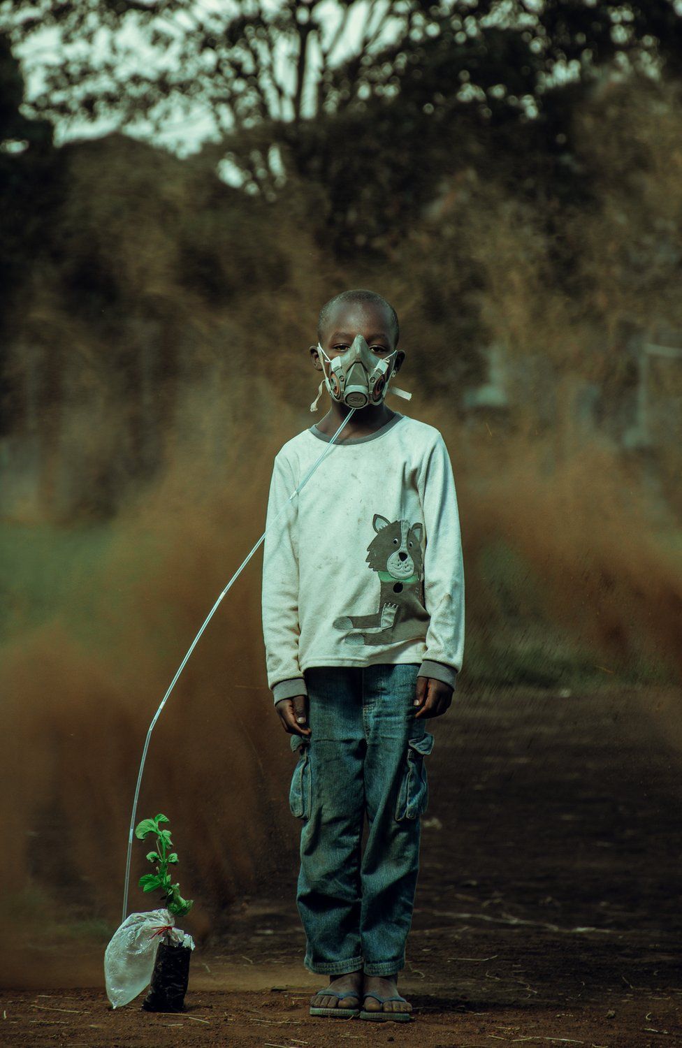 A boy is seen wearing an oxygen mask attached to a plant, with a sand storm behind him