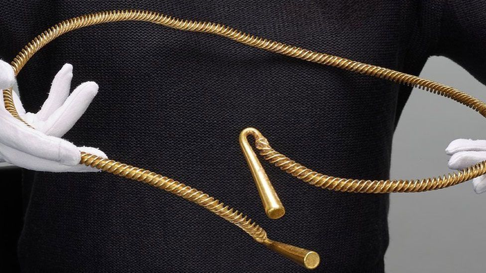 A large Bronze Age torc made of twisted strands of gold held by white gloved hands in front of a black-clad torso