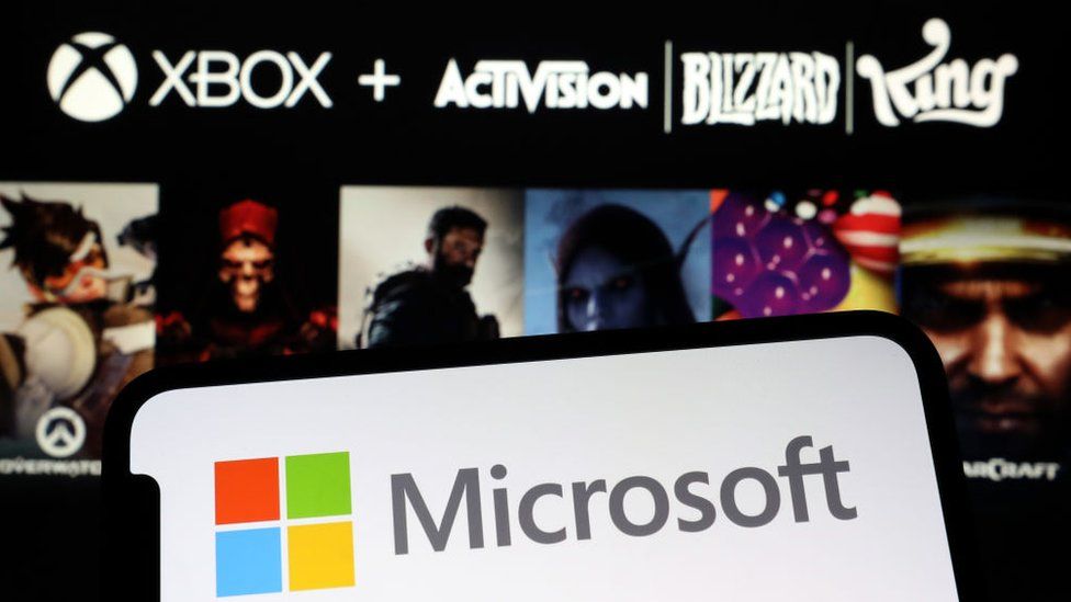 Смартфон показывает логотип Microsoft в виде красного, зеленого, желтого и синего квадрата напротив названия компании на пустом экране. Фон размыт, но видны очертания персонажей из известных игр Blizzard и Activision. Над ними видны логотипы компаний Xbox, Activision, Blizzard и King