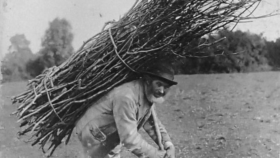 Black and white photo of a thatcher. He has a grey beard and weathered face and is stooping. His hands hold a pole supporting a bundle of hazel on his back.