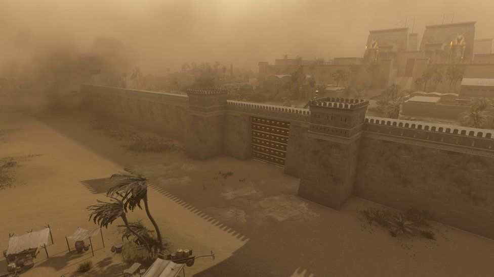 The fortified walls of an Ancient Egyptian town are swathed in clouds of brown sand, creating a foggy, limited field of vision across the scene. We can see a heavy gate with a square guard towers built into the wall at either side. We can just make out various buildings within the city walls, most are quite low with flat or white domed roofs. At the rear of the scene, two larger towers, with ornate statues of a half-man, half-goat Egyptian god standing in front of each, can be seen.