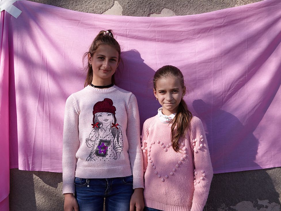 Two girls from a Ukrainian village pose against a pink cloth backdrop