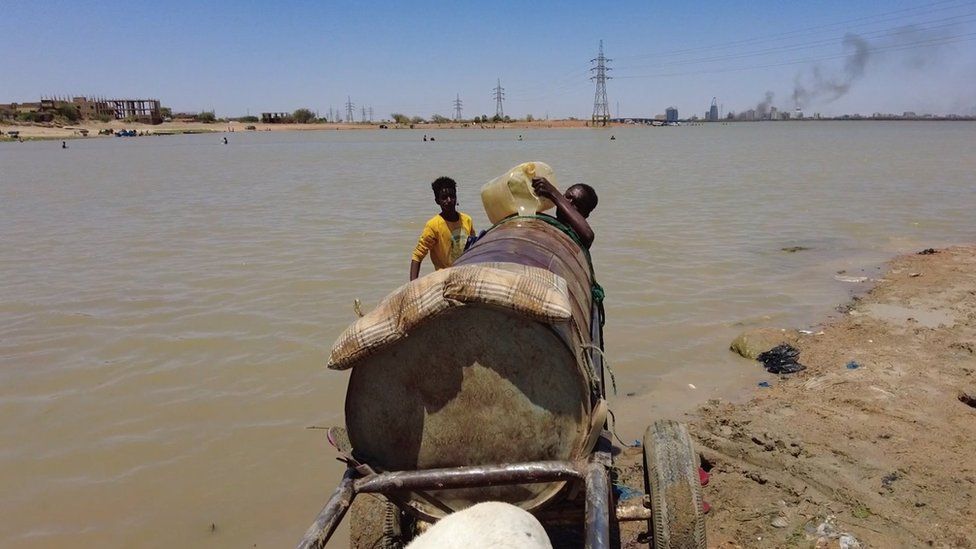 Two boys fill drum up on a cart, with water from buckets from the river