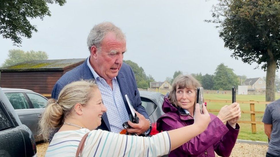 Jeremy Clarkson with two women who are holding up their phones and posing for selfies