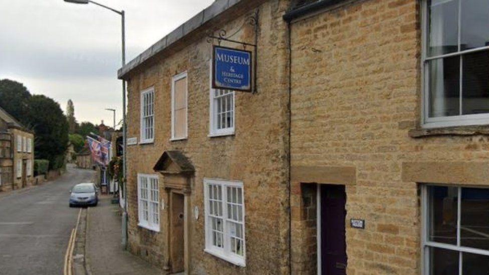 The front of Crewkerne and District Museum and Heritage Centre, which is an old listed building with a blue pub-style sign along a road in the town