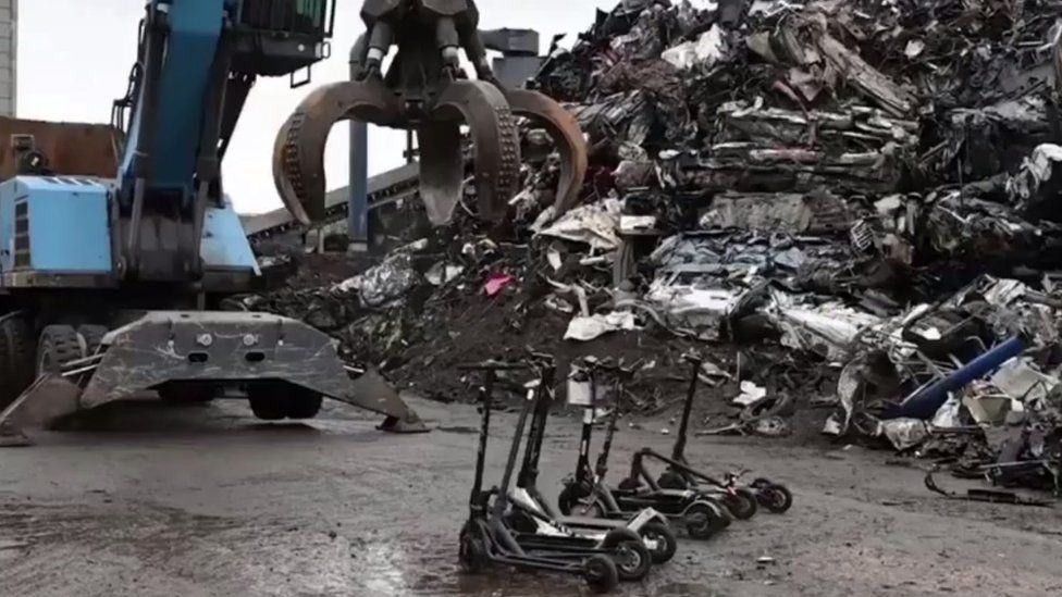 E-scooters lined up to be grabbed by a blue mechanical claw next to a pile of scrap metal
