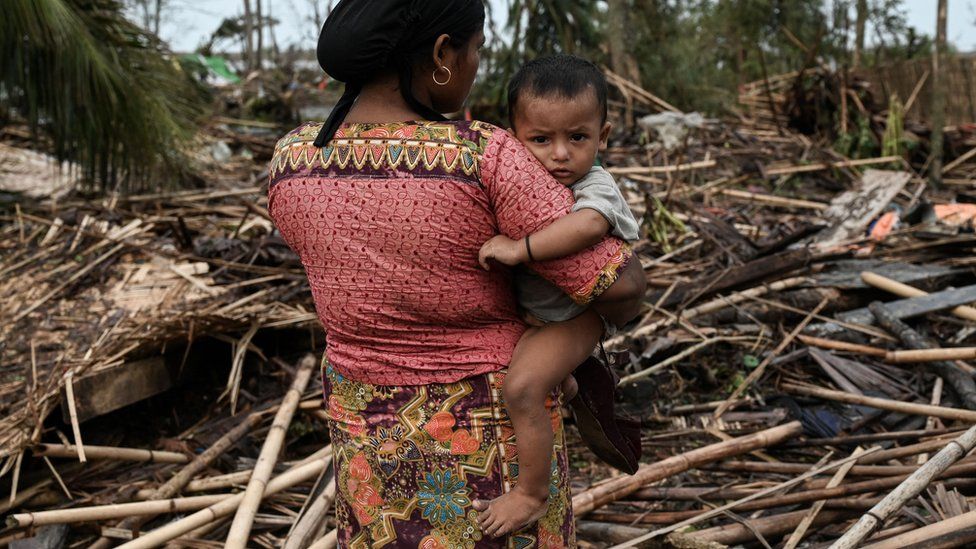 A Rohingya woman carries her baby next to her destroyed house at refugee camp in Sittwe, Myanmar. Photo: 16 May 2023