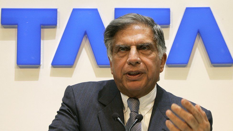Ratan Tata sitting in front of a Tata sign