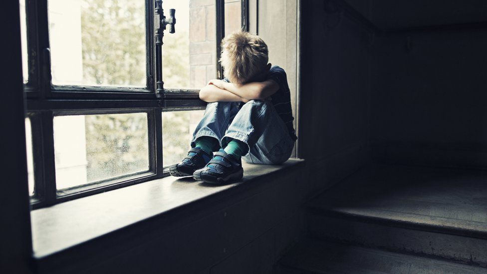 A child sits hunched on a windowsill, head in his arms, crying