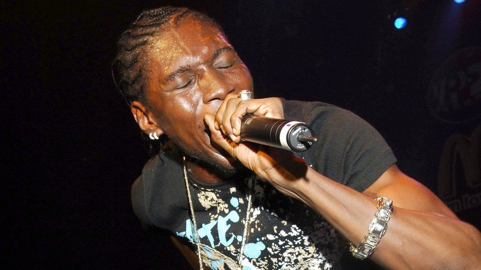 A black man with short cornrows holds a microphone to his lips as he performs on stage. His eyes are closed, suggesting a passionate performance. He wears a large ring on one finger, has two hoop earrings in one ear, and wears a long silver chain over a black t-shirt with an elaborate, colourful design. The arm holding the mic has a large, chunky silver bangle around the wrist.