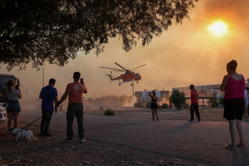 People look at a firefighting helicopter filling water from a pool, as a wildfire burns in the village of Gennadi, on Rhodes