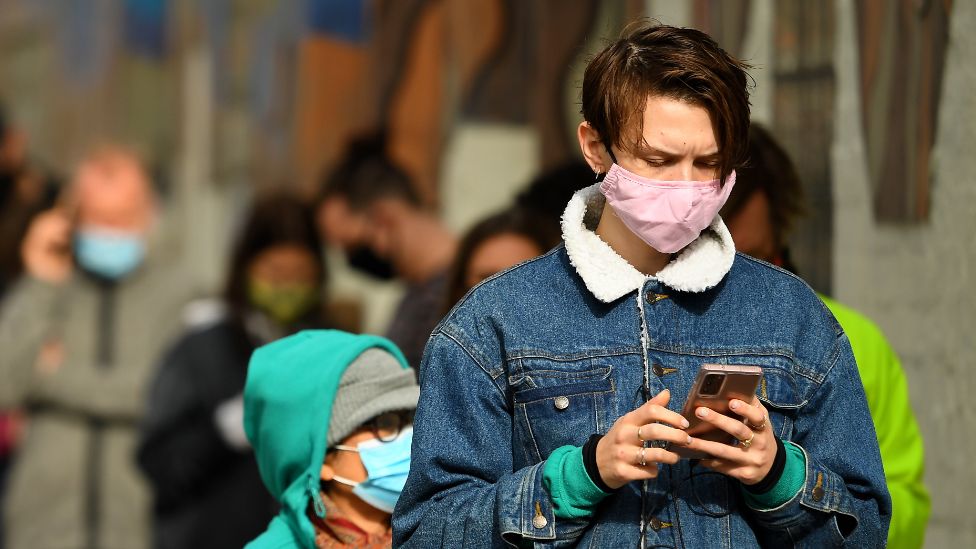 A young man wearing a mask checks his phone while queueing for a Covid test in Melbourne