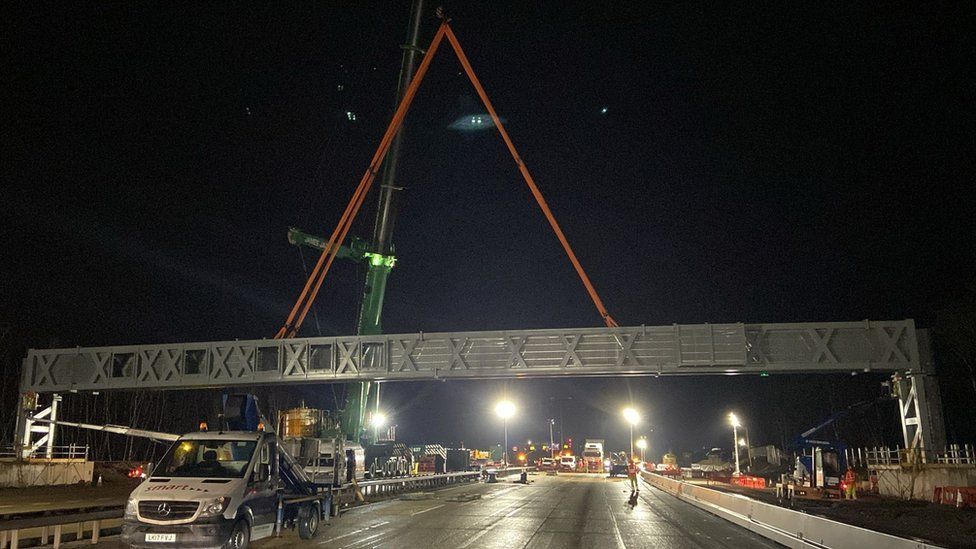 A night time image shows a gantry being lifted into place above the M25