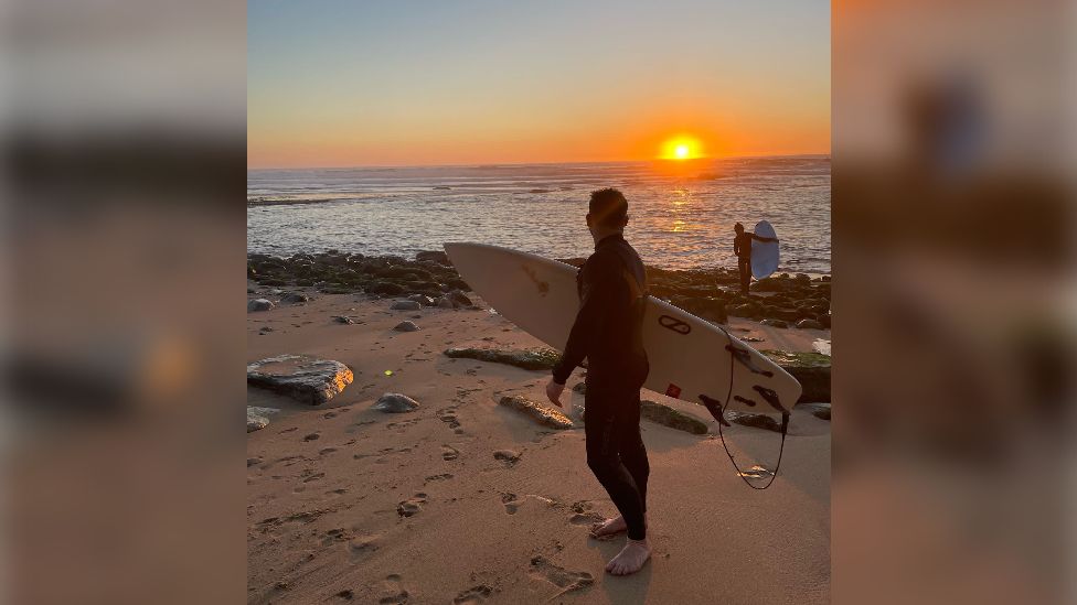 Dan Richards on the beach holding a surfboard with the sun setting