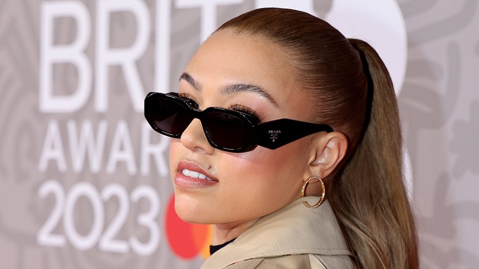 Mahalia looks over her shoulder at the camera while wearing sunglasses, with the Brit Awards 2023 logo behind her
