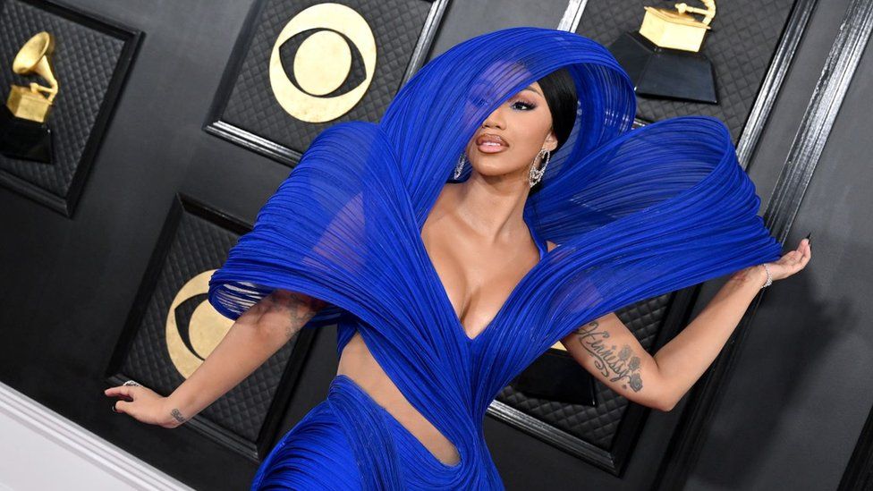 Cardi B attends the 65th GRAMMY Awards in LA. Cardi wears one of Gaurav's designs, a swirling royal blue dress with exaggerated shoulders and a hood partially covering her face. She has her black hair tied back and wears dangling silver earrings. Cardi is pictured on the red carpet in front of a black and gold backdrop
