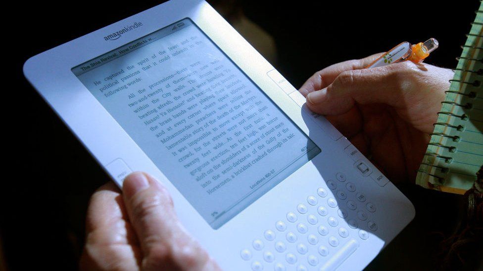 A white Amazon Kindle 2 is held by a person next to a pen and notepad