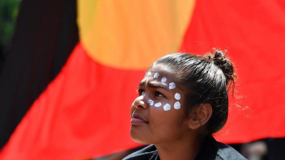 A young woman with ceremonial face paint in front of the Aboriginal flag