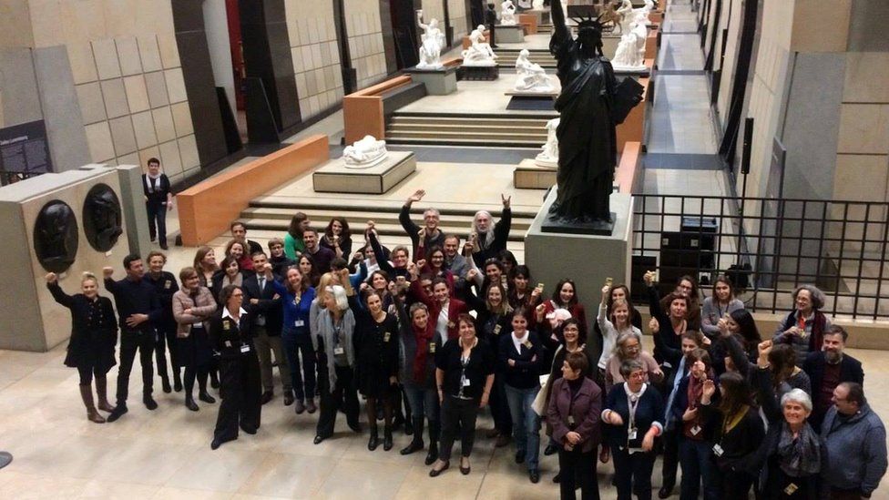 Group of female and male employees in the atrium of musee d'orsay raise hands in show of solidarity