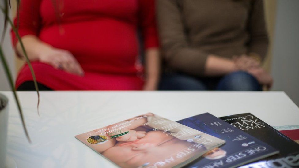 Two surrogates, sit in a waiting room in front of a coffee table with magazines, one of which has a baby's face on the cover