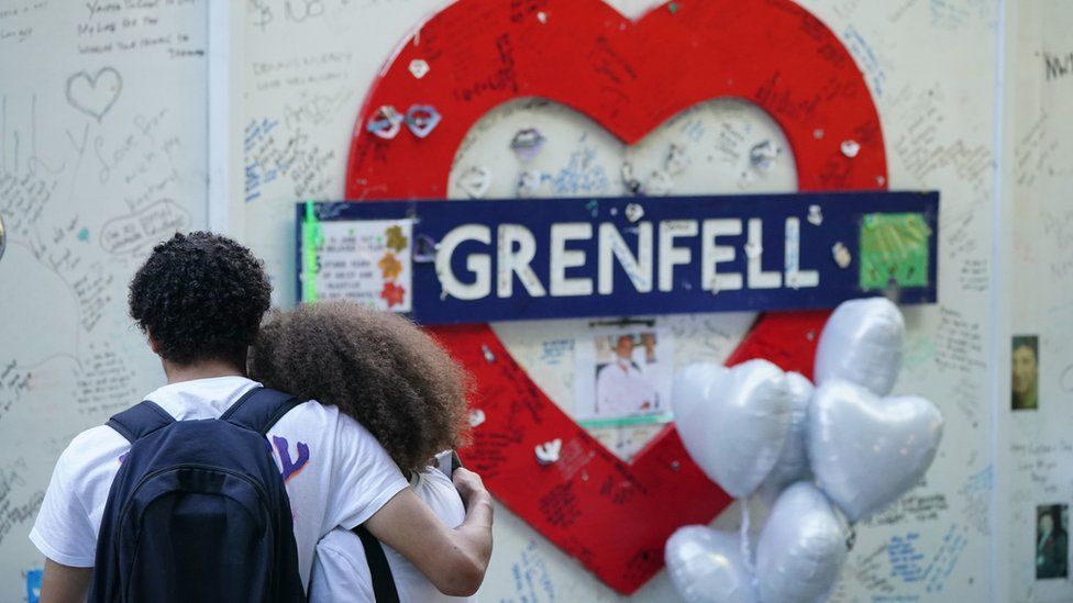 Two people hug in front of a sign for Grenfell