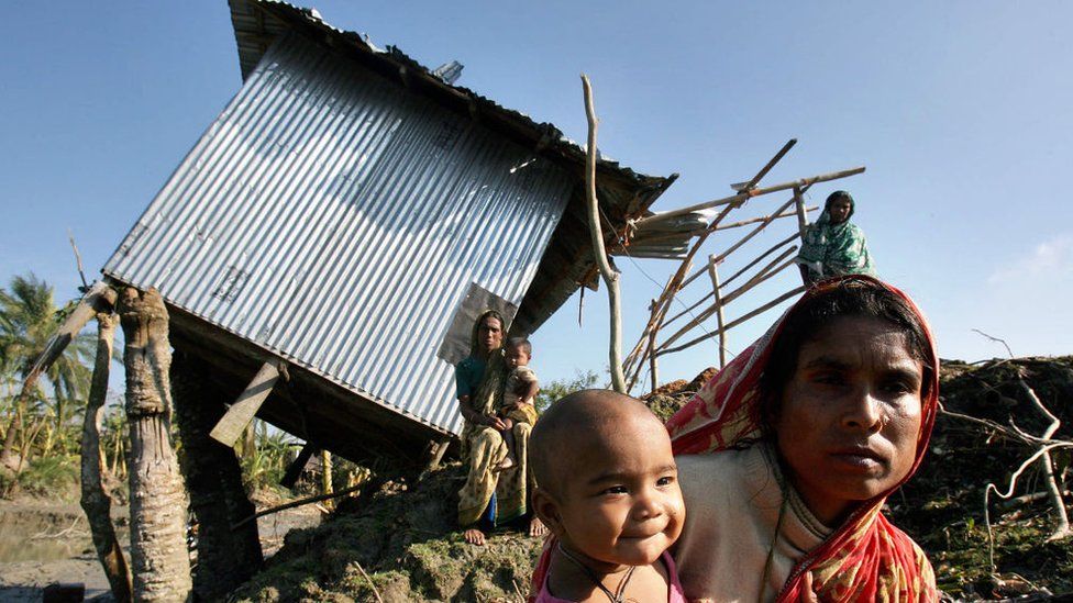 Women and children stand in front of a metal shack tilted on to its side