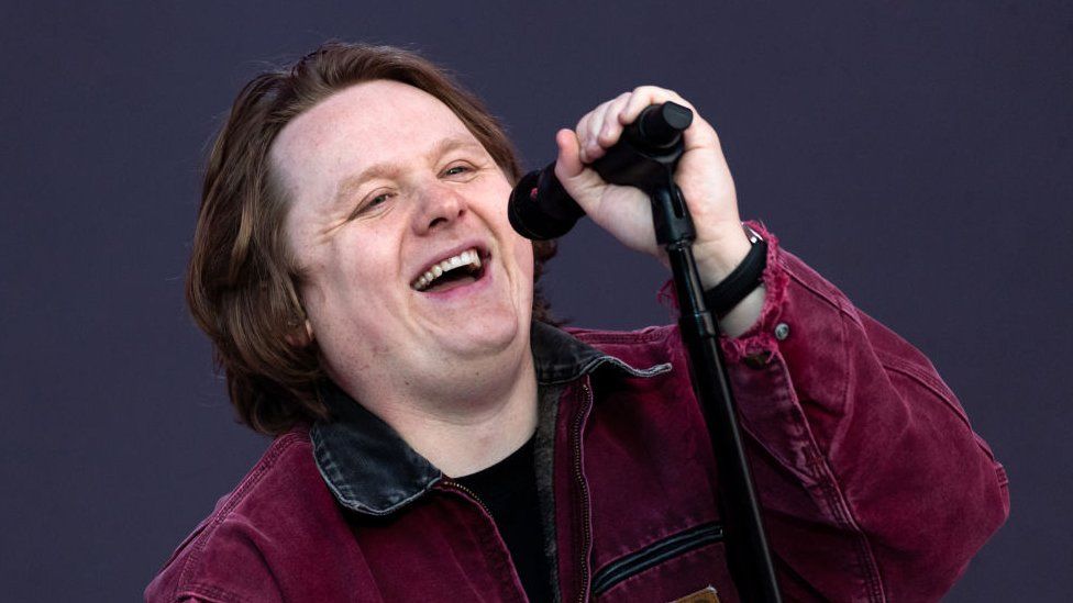 Lewis Capaldi on stage during Radio 1's Big Weekend in Dundee in 2023. Lewis is a 27-year-old white man with brown hair almost to his shoulders. He wears a burgundy jacket with a black collar and smiles as he sings into a microphone, which he holds toward him with his left hand. He is pictured on stage in front of a dark grey backdrop