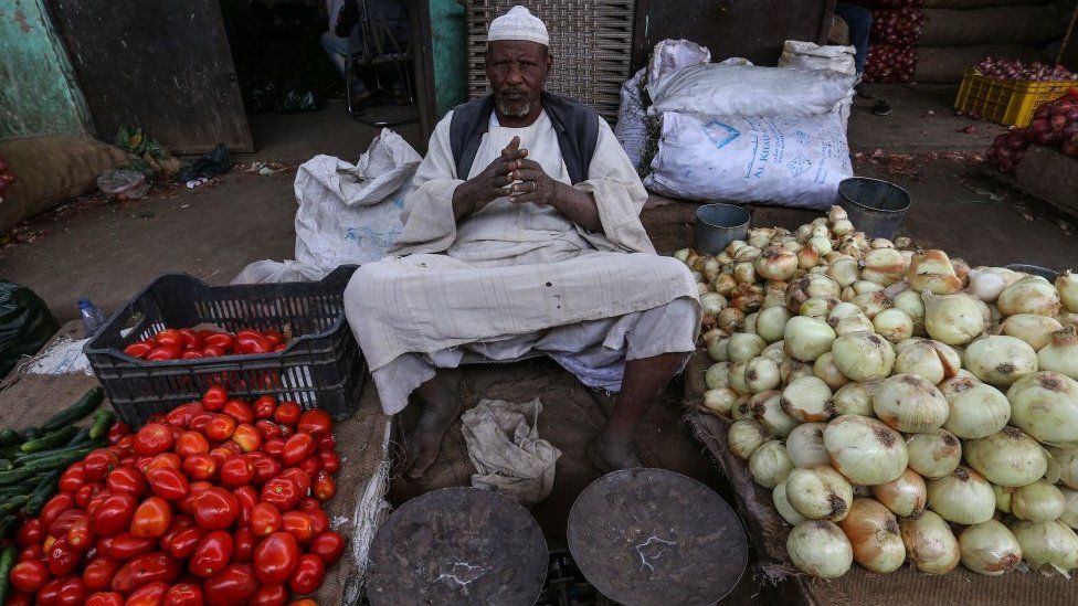 A market seller in Sudan sitting down. There are vegetables to his left and right.