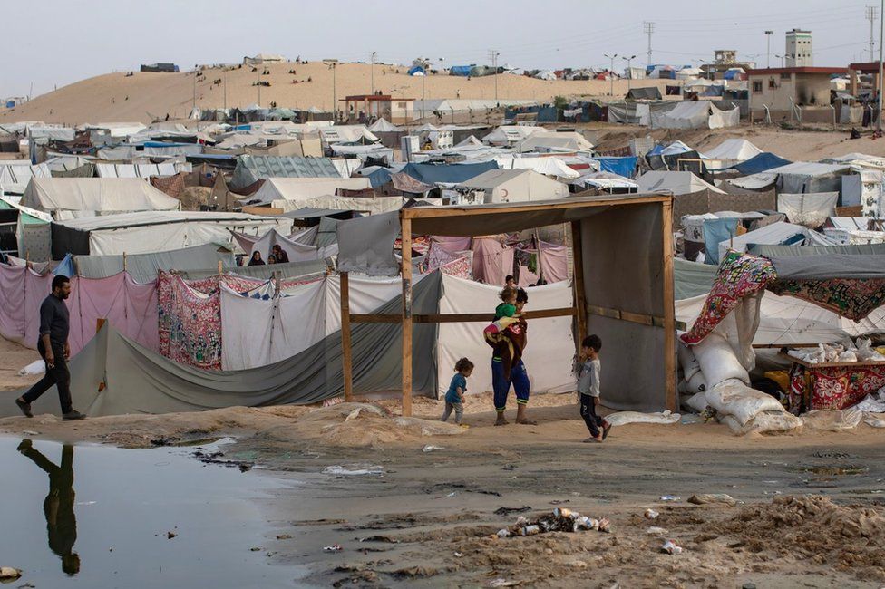 Palestinians walk past a pool of sewage at a temporary camp for displaced people in Rafah, in the southern Gaza Strip (26 April 2024)