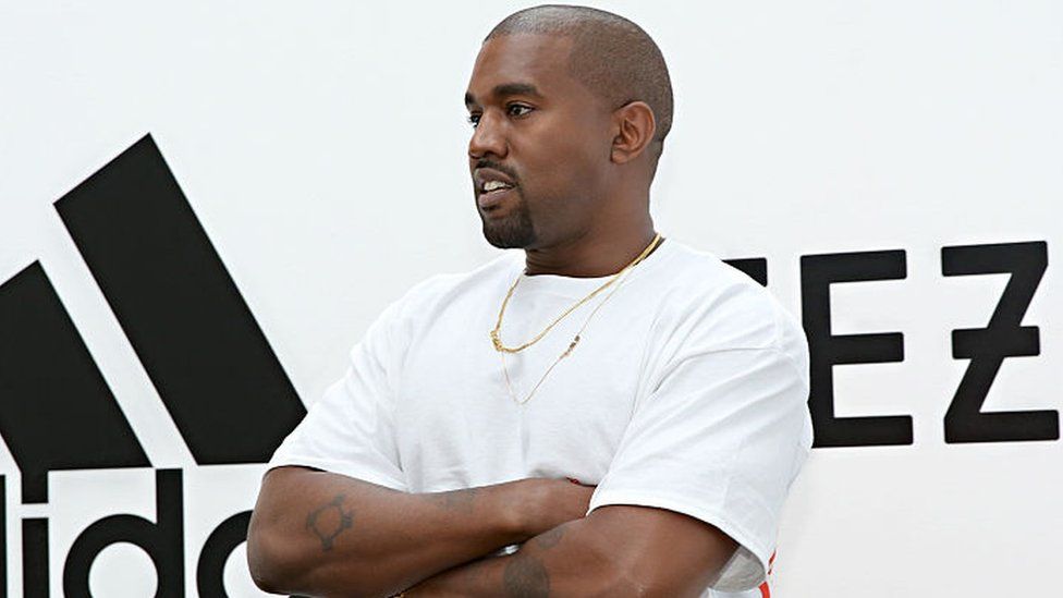 Kanye West in front of Adidas and Yeezy logos.
