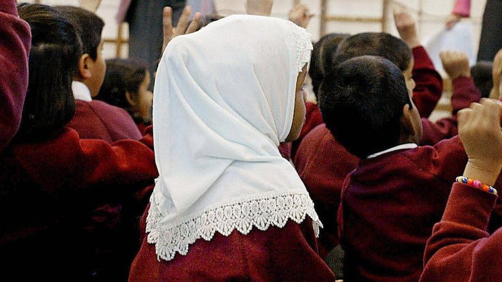 A young girl wearing a Muslim headscarf listens as two teachers instruct a class.