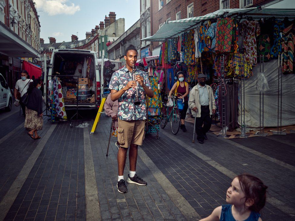 A musician stands on a busy market street in Brixton, London, playing a flute