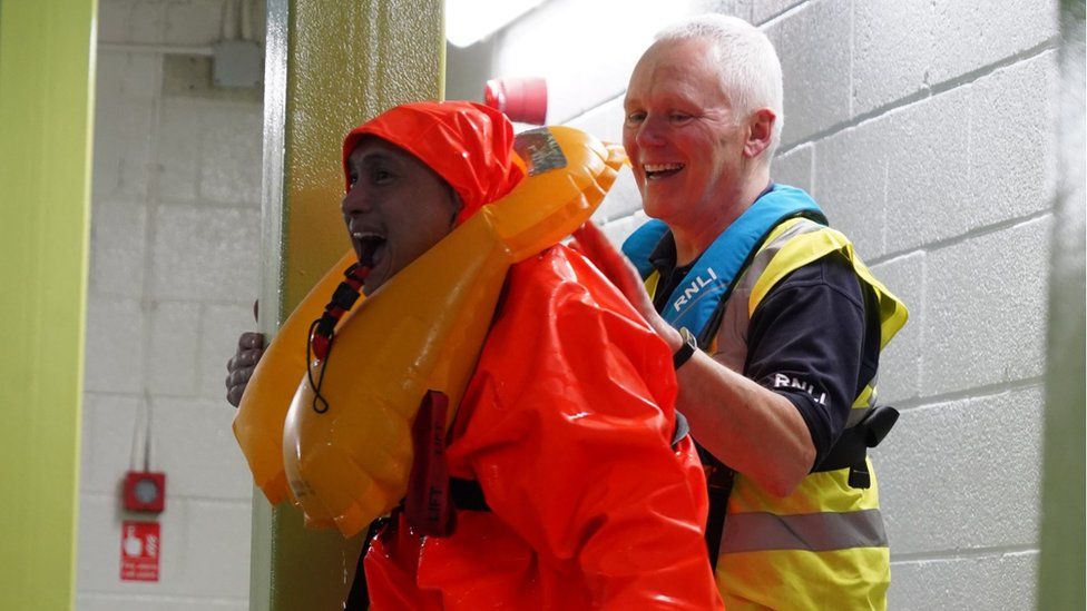 A man in a large water proof suit is held up by Geoff beside a pool