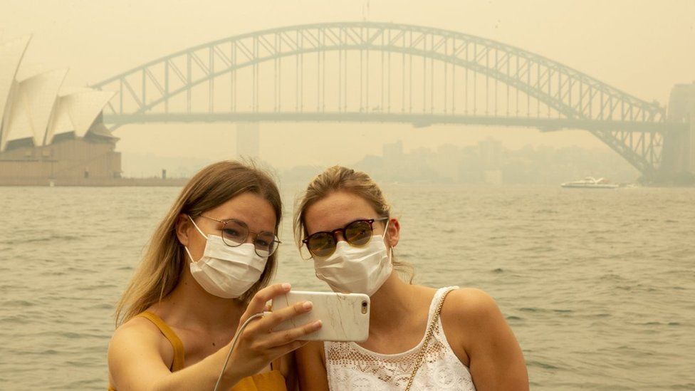 Two German tourists pose for a selfie, wearing breathing masks, as heavy smoke blankets Sydney skyline