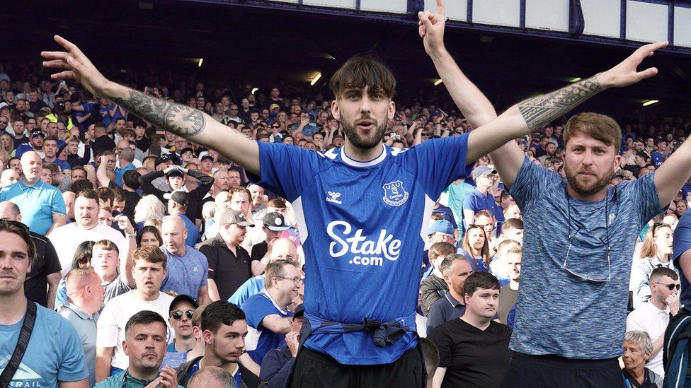Two Everton fans in the stands at a game, both have their arms raised up in a celebratory pose. One wears a blue Everton home shirt, with the club's badge over the left breast and the Stake . com logo in the centre.