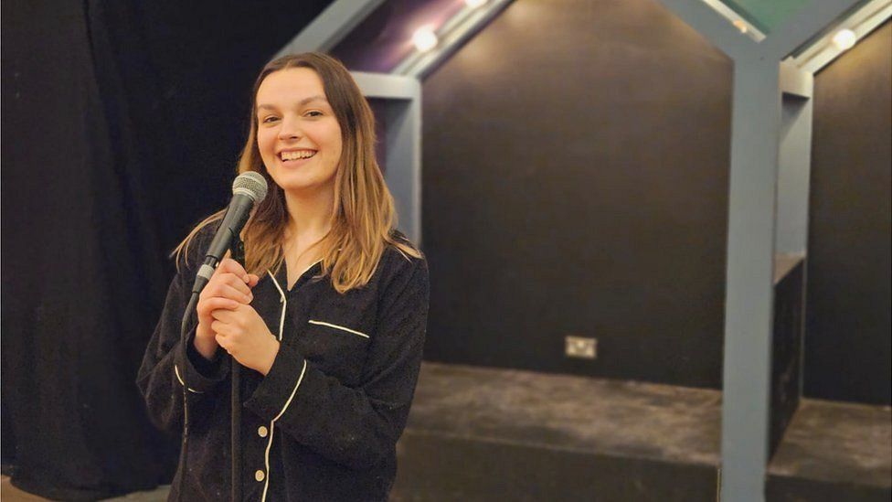 Millie Malone holding a microphone and smiling, wearing black pyjamas