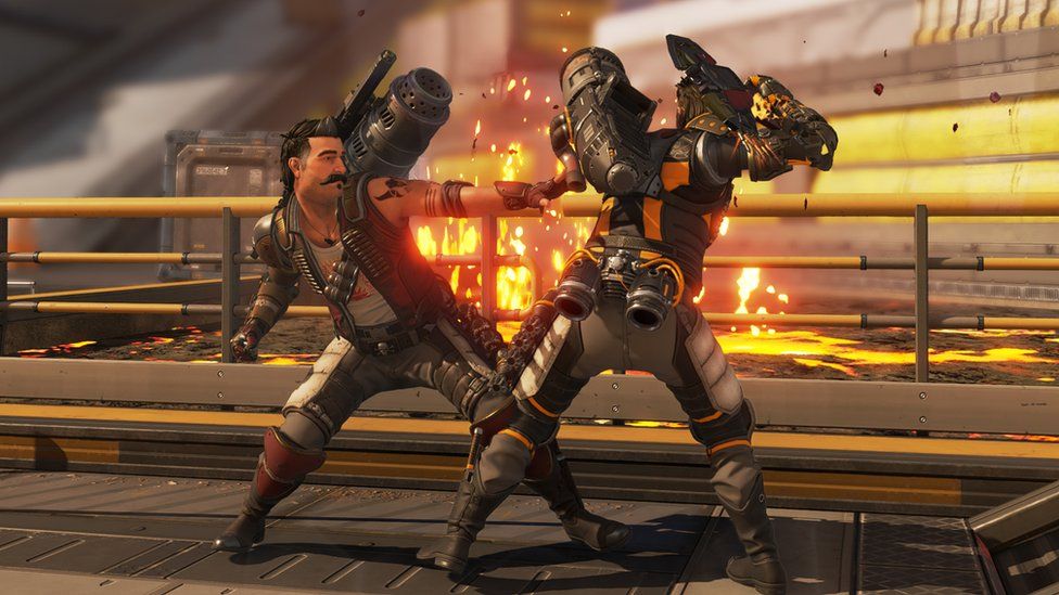 Two characters battle hand-to-hand in an industrial, factory-style environment on a metal walkway. One is facing the viewer, the other has their back to the camera. Both have large, tubular rocket-launcher weapons attached to their shoulders and have ammo belts and grenade holders attached to their bodies. A fire burns in the background as one character lands a punch on his opponent.