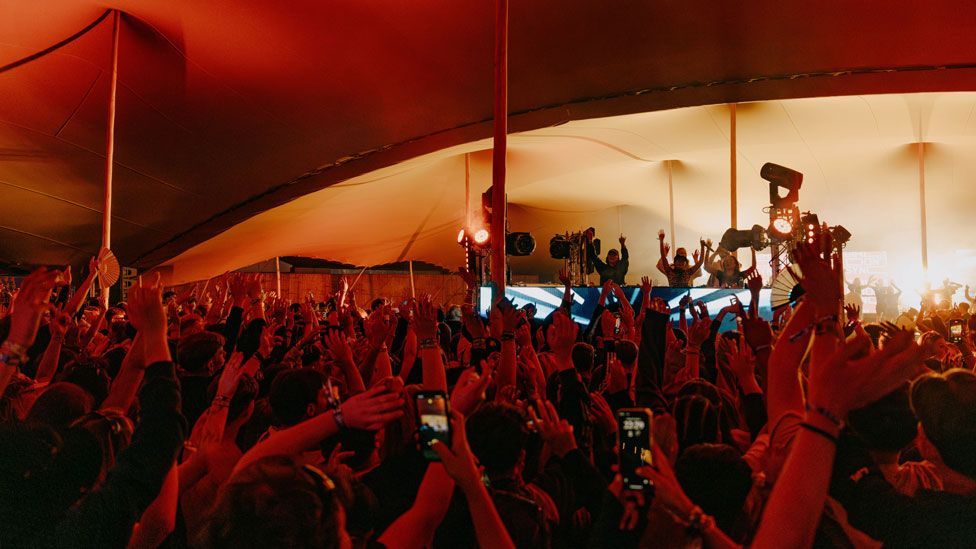 Hundreds of people with their hands raised some with phones held up in front of a band on a stage in a deep red light