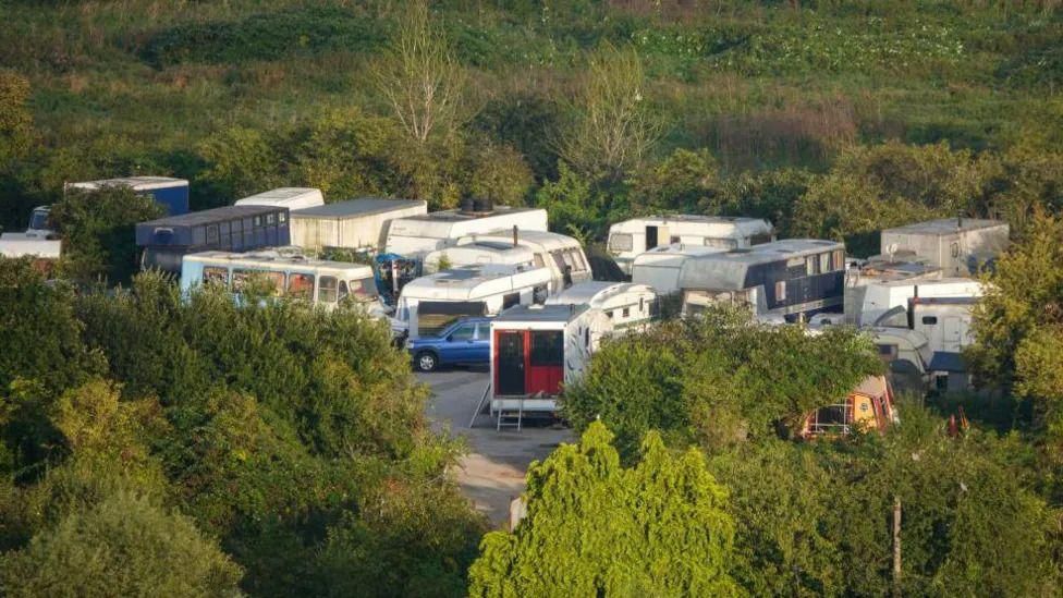 Roughly a dozen caravans and vehicles parked in a car park. The car park is surrounded by green fields and trees.