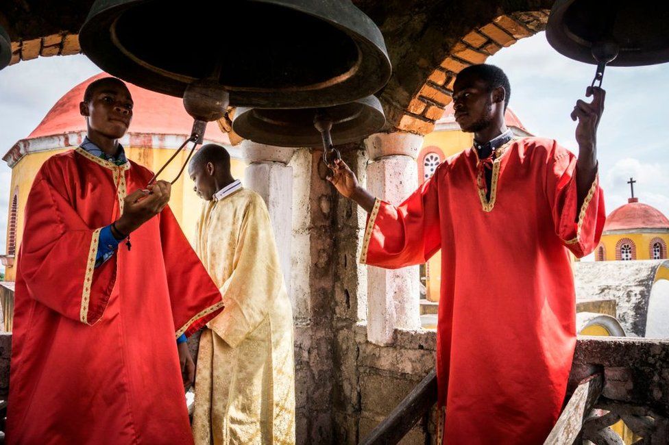 Men dressed in red clerical robes ring large church bells