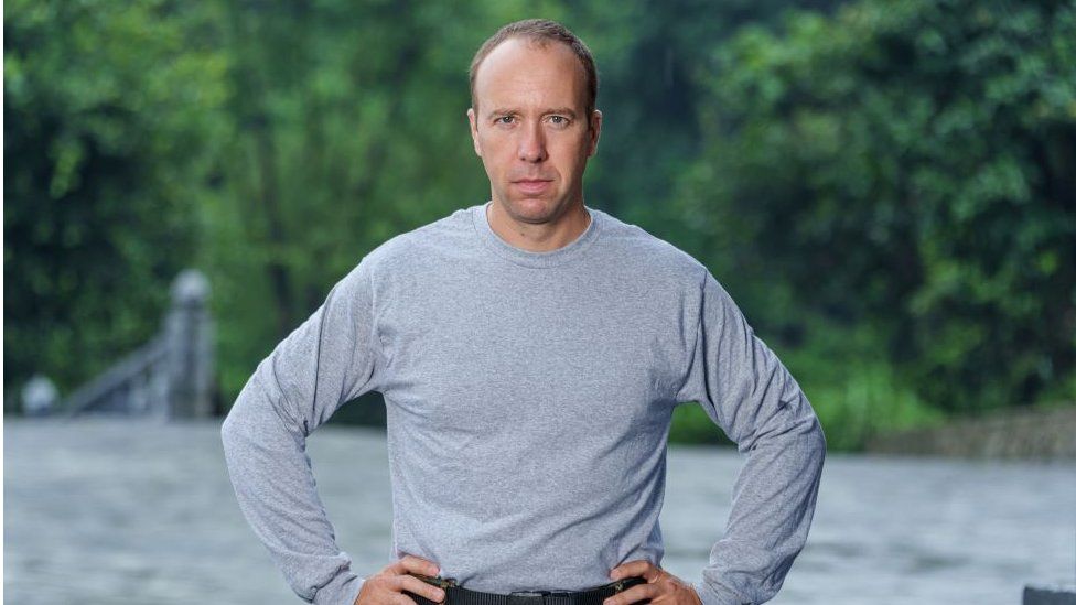 Matt Hancock wearing a grey t-shirt and green army cargo pants. He is standing in front of a river with his hands on his hips.